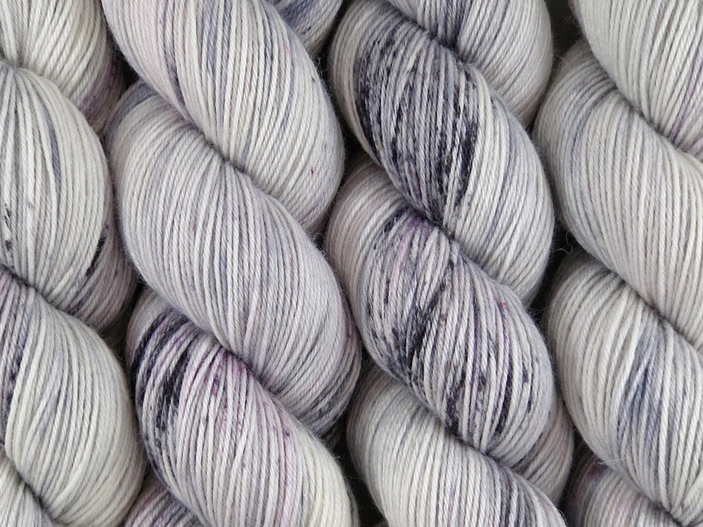 A close up of white and soft grey with speckles of black and purple coloured skeins of superwash merino and nylon 4ply fingering sock yarn (Newsprint on Tough Stocking)