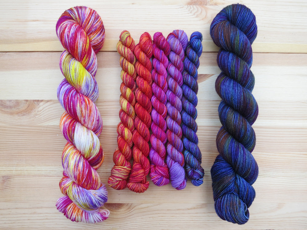 Five brightly coloured variegated mini skeins of yarn flanked by two full sized skeins lined up vertically on a pale wooden background