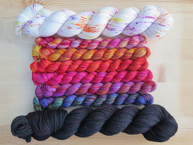 Six brightly coloured variegated mini skeins of yarn flanked by two full sized skeins lined up horizontally on a pale wooden background