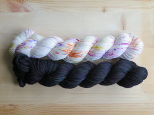 A white skein of yarn with bright coloured speckles and a black skein of yarn with variegated undertones   lie horizontally on a pale wooden background