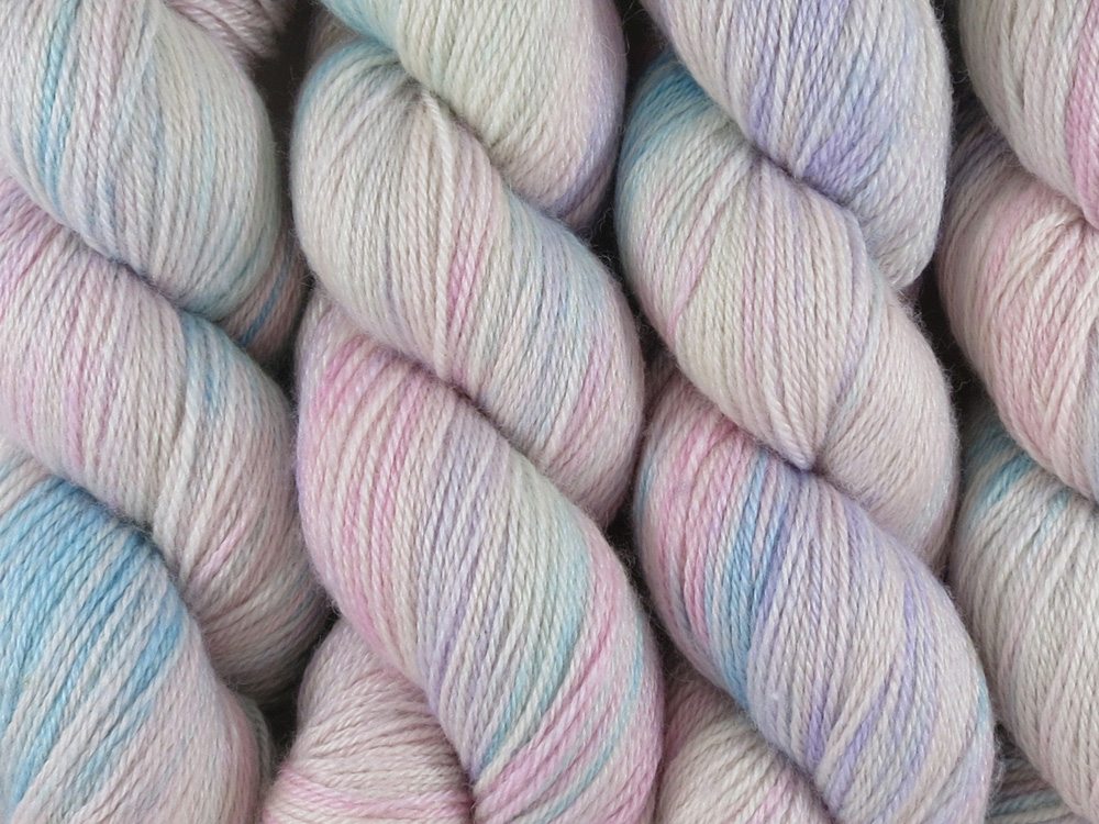 A close up of variegated white, soft pink, blue and violet coloured skeins of superwash bluefaced leicester, silk and cashmere 4ply fingering sock yarn (White Opal on Blue Chip Stocking)