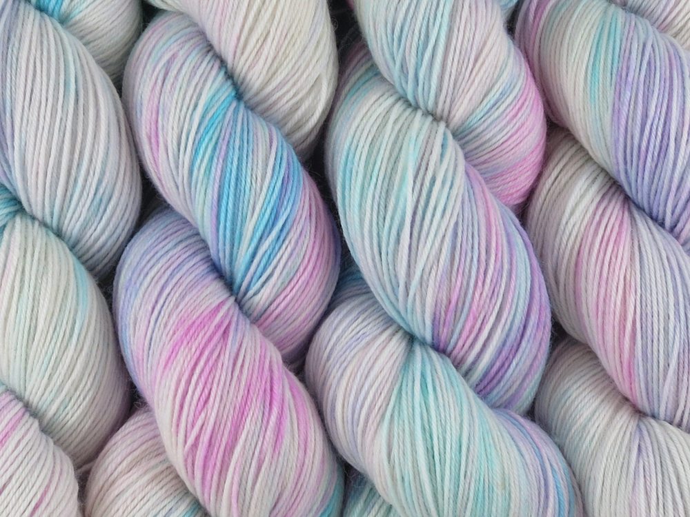 A close up of variegated white, soft pink, blue and violet coloured skeins of superwash merino and nylon 4ply fingering sock yarn (White Opal on Tough Stocking)