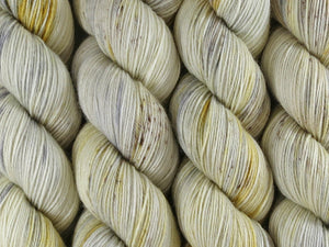 A close up of variegated warm white, golden yellow, grey with brown speckles coloured skeins of superwash merino and nylon 4ply fingering sock yarn (Witchetty Grub on Tough Stocking)
